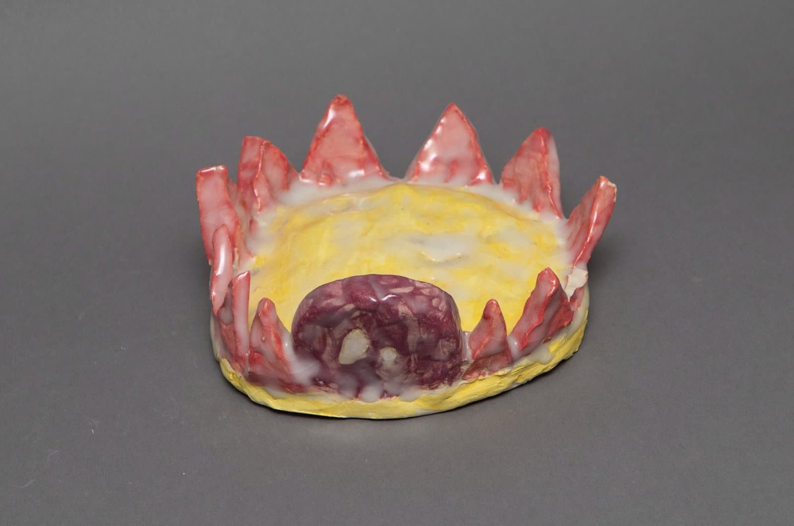 Ceramic sculpture resembling a crown with pointed red spikes and yellow base containing a dark purple central form.