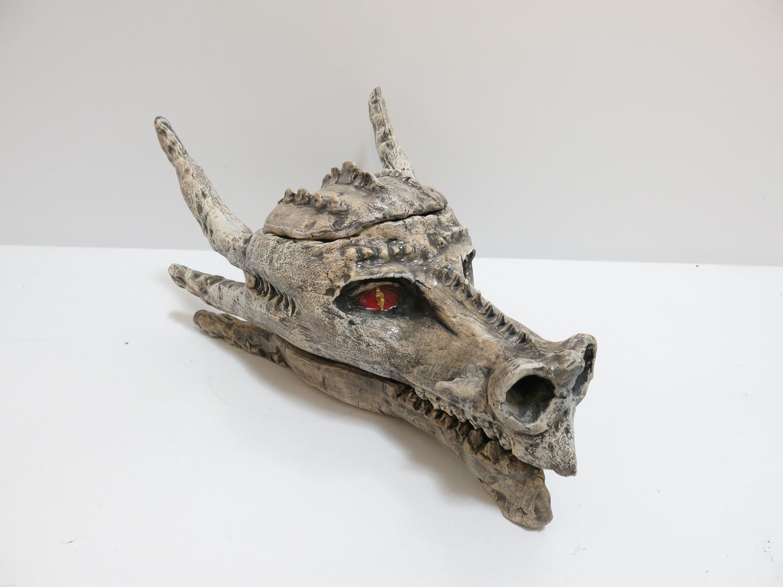 Ceramic sculpture of a dragon skull with horns and a single red eye, finished in mottled gray-white glaze.