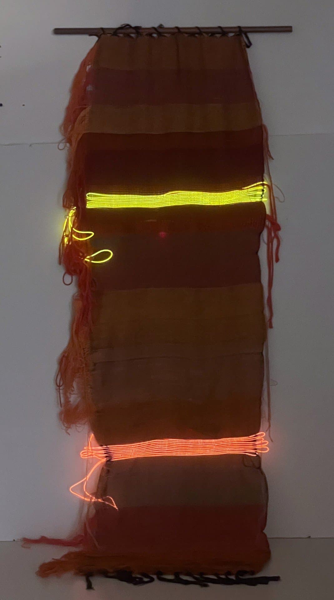 Vertical textile weaving in earth tones with integrated yellow and orange neon lighting, mounted on wooden rod.