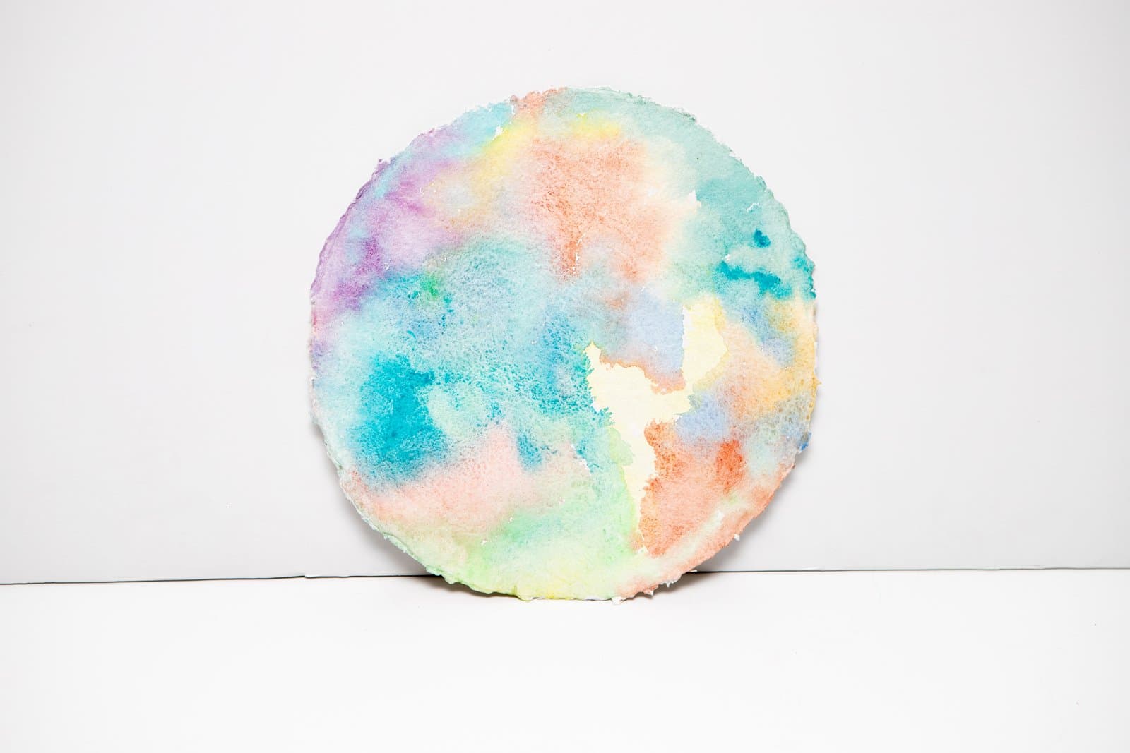 Circular watercolor painting with soft, blended pastel washes of teal, pink, lavender, yellow, and orange on white paper.