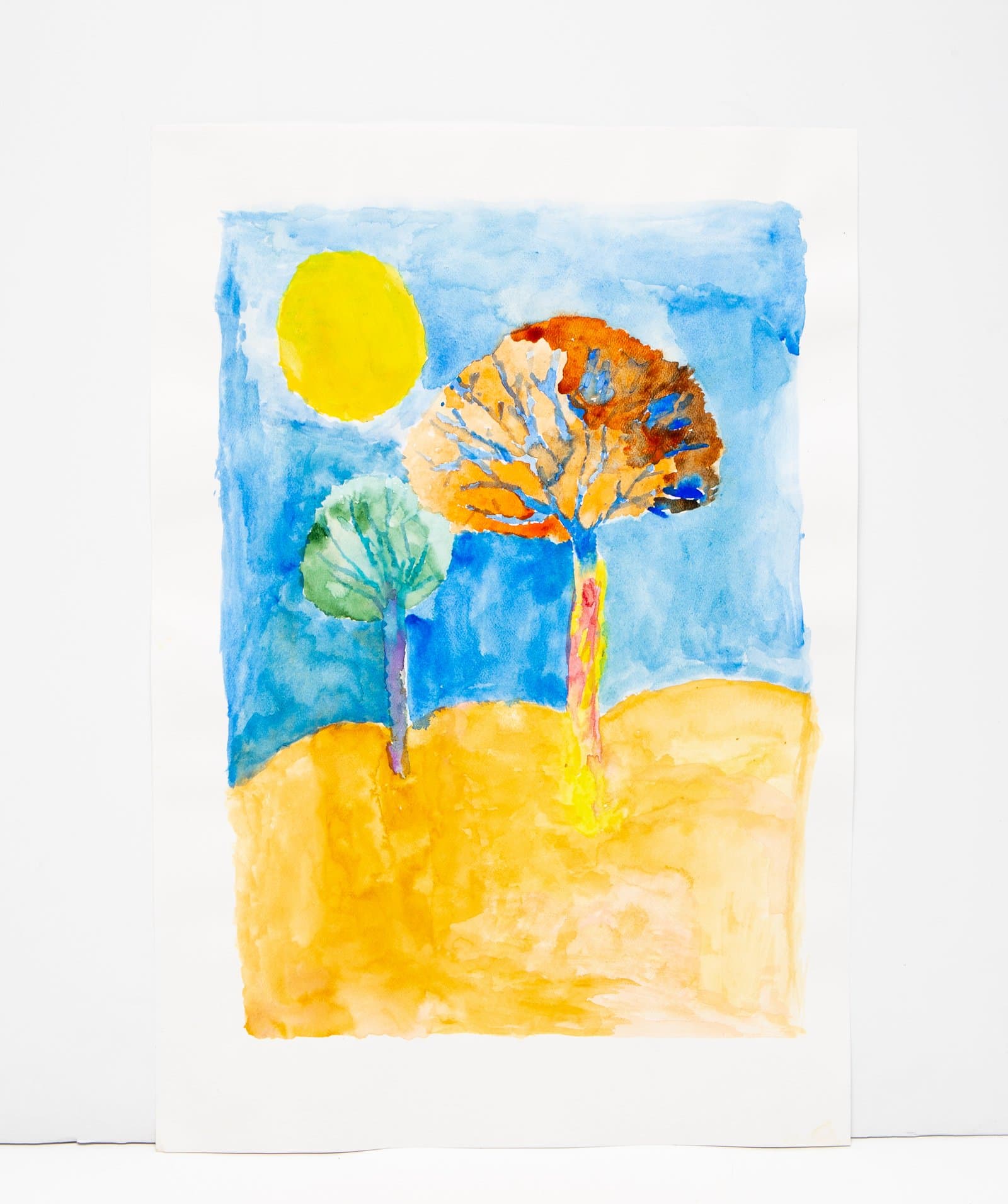 Watercolor landscape with two trees, a yellow sun, blue sky, and golden ground in warm and cool tones.