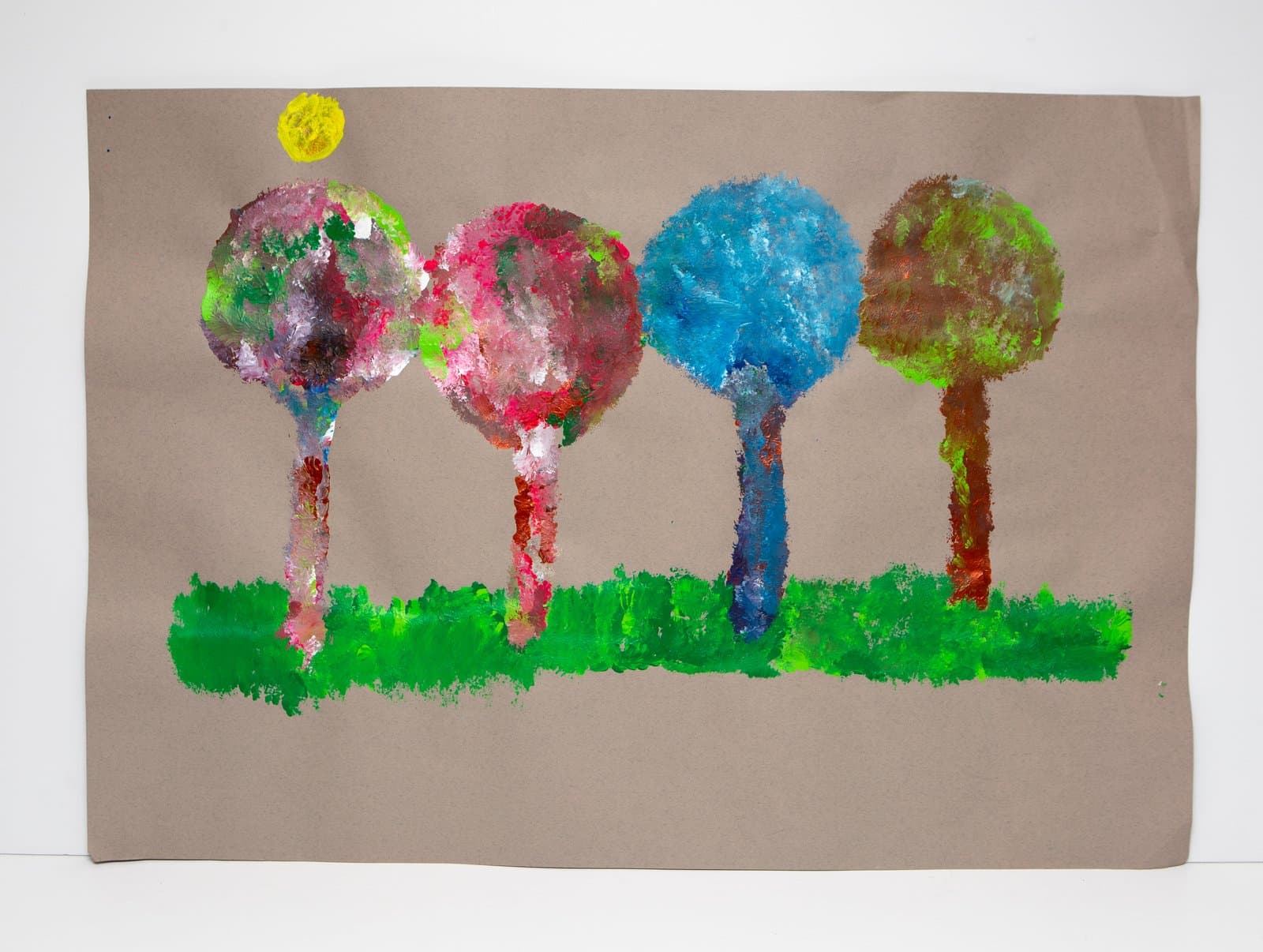 Acrylic painting of four round-topped trees and a yellow sun on brown paper with a bright green ground line.