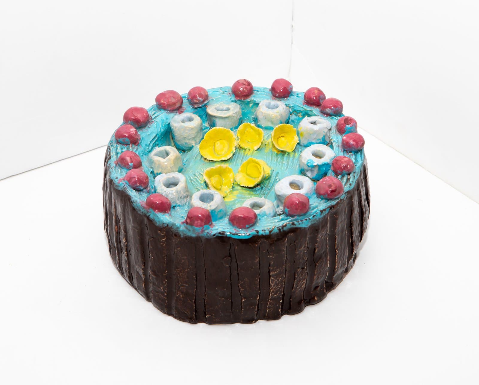 Ceramic sculpture resembling a round cake with dark brown sides, turquoise top, and decorative elements in red, white, and yellow.