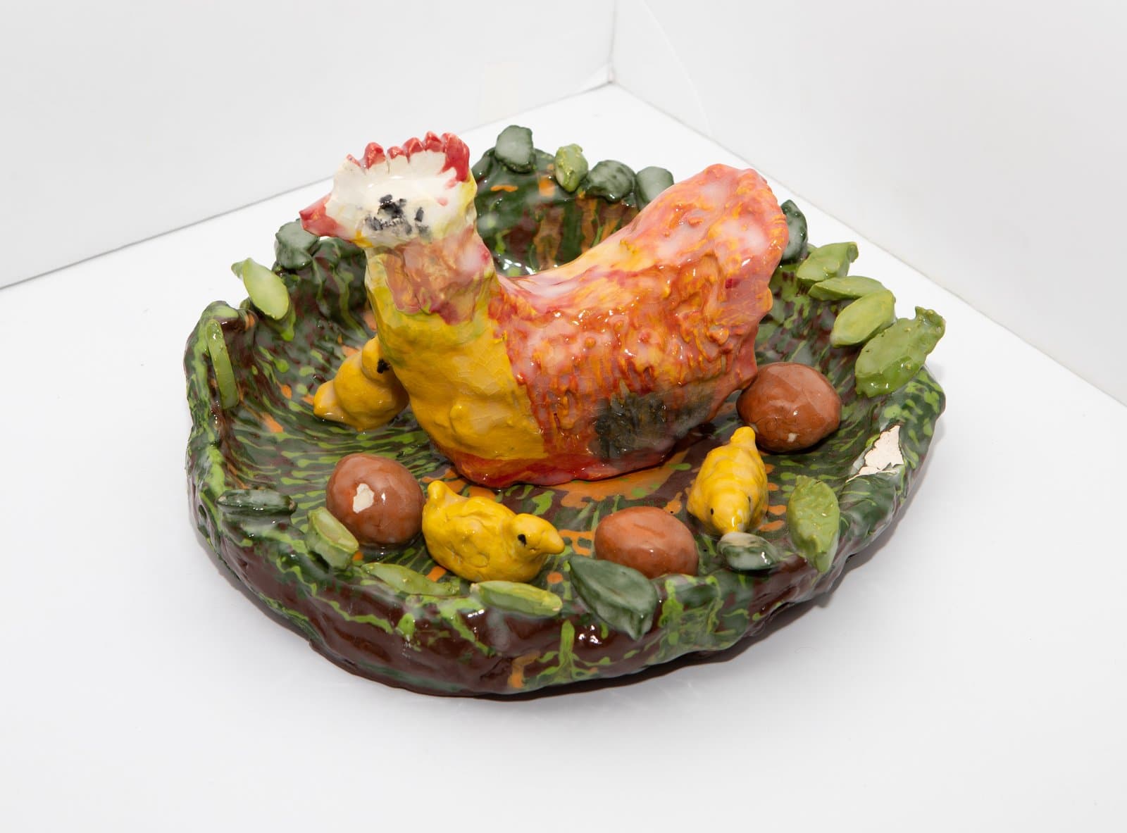 Glazed ceramic sculpture of a hen on a nest-like dish with chicks, eggs, and foliage in red, yellow, and green.