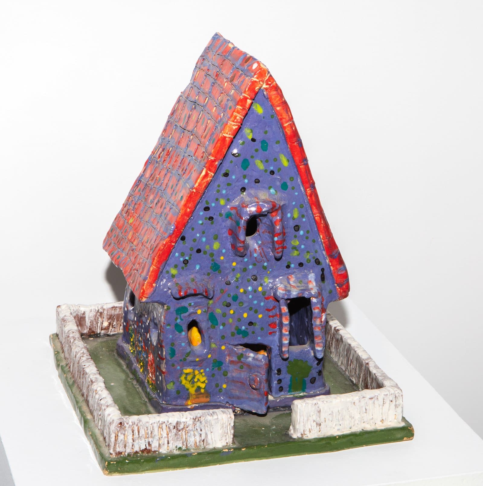 Ceramic sculpture of a house with a steep red grid-patterned roof, purple walls with multicolored dots, and a white fence base.