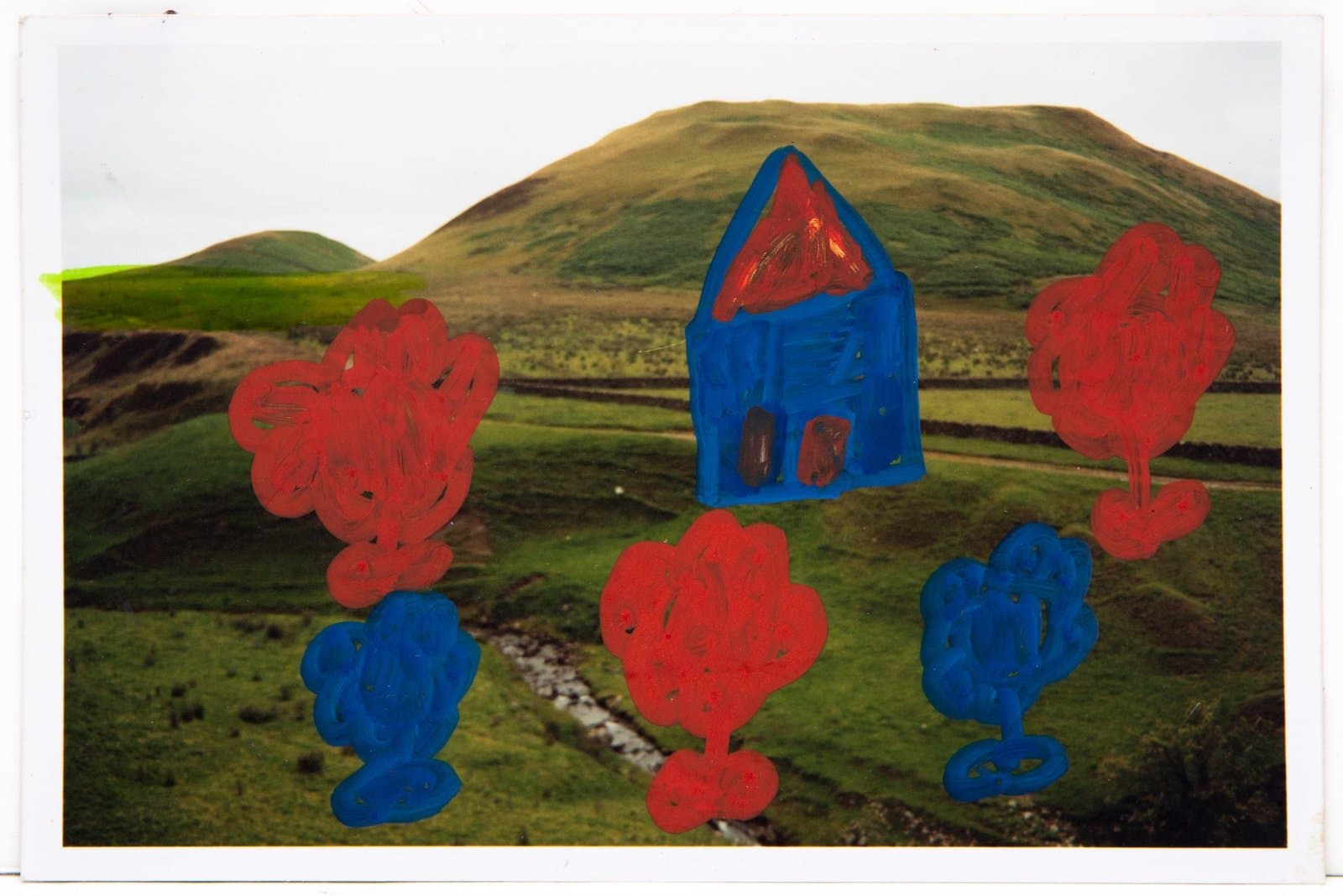 Mixed media artwork with blue house and red and blue trees painted over a landscape photograph of rolling hills