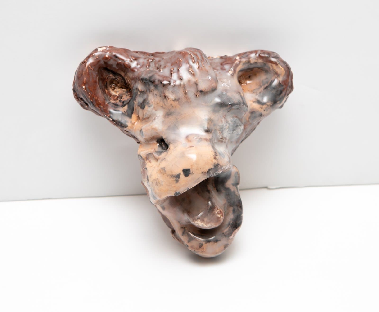 Small glazed ceramic sculpture of an animal head with open mouth, in mottled peach, brown, and gray tones.