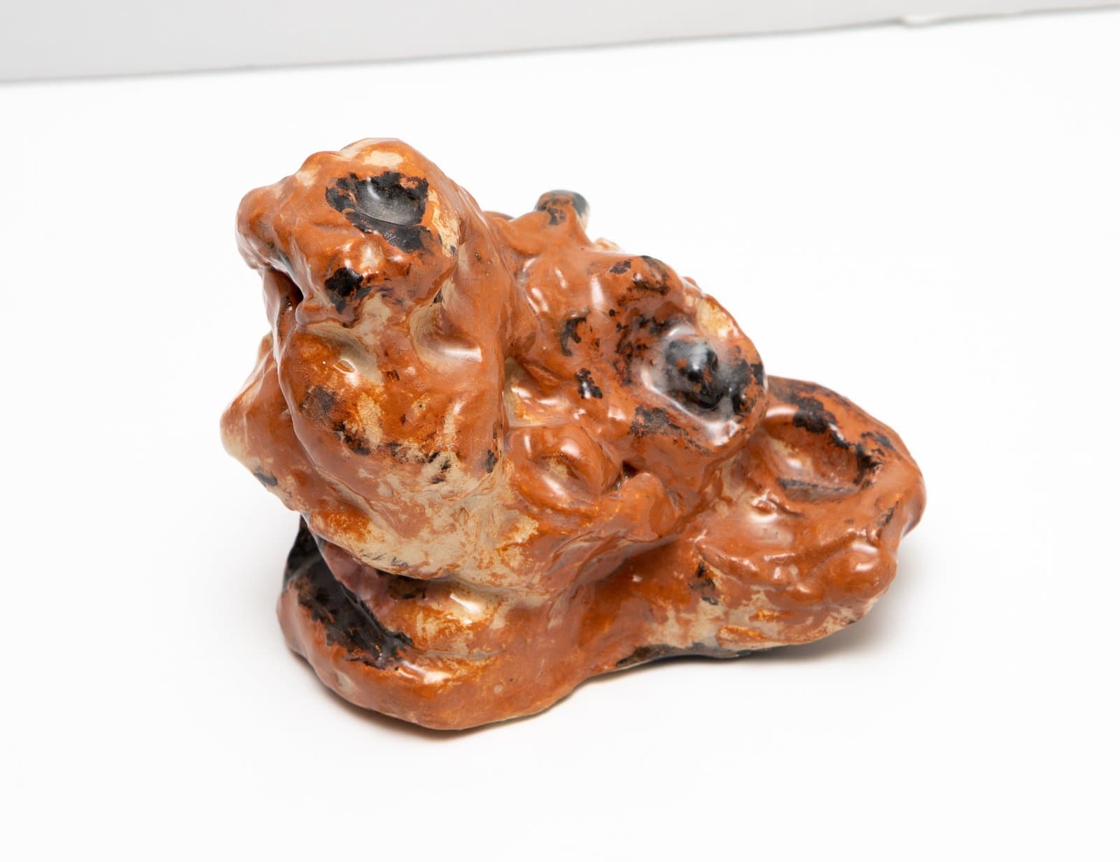 Small glazed ceramic sculpture with lumpy, organic form in terracotta orange with black glaze patches, on white surface.