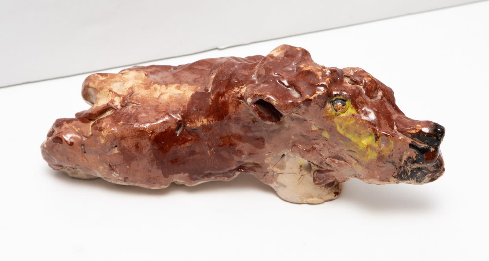 Glazed ceramic sculpture of a bear in profile, low and horizontal, with brown glaze and yellow accents near the face.