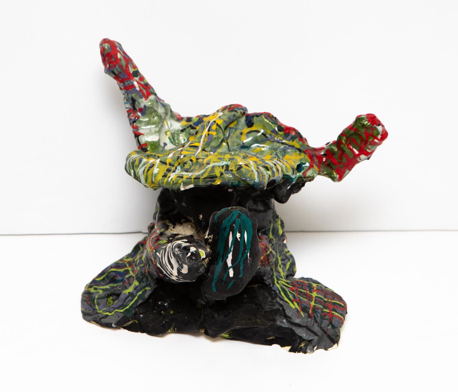 Ceramic sculpture with a black textured base and a wide, irregular painted top in green, red, and yellow with two horn-like protrusions.