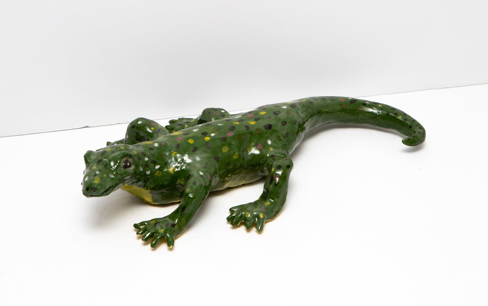 Ceramic sculpture of a lizard in glossy dark green glaze with multicolored spots, posed in a crawling position.