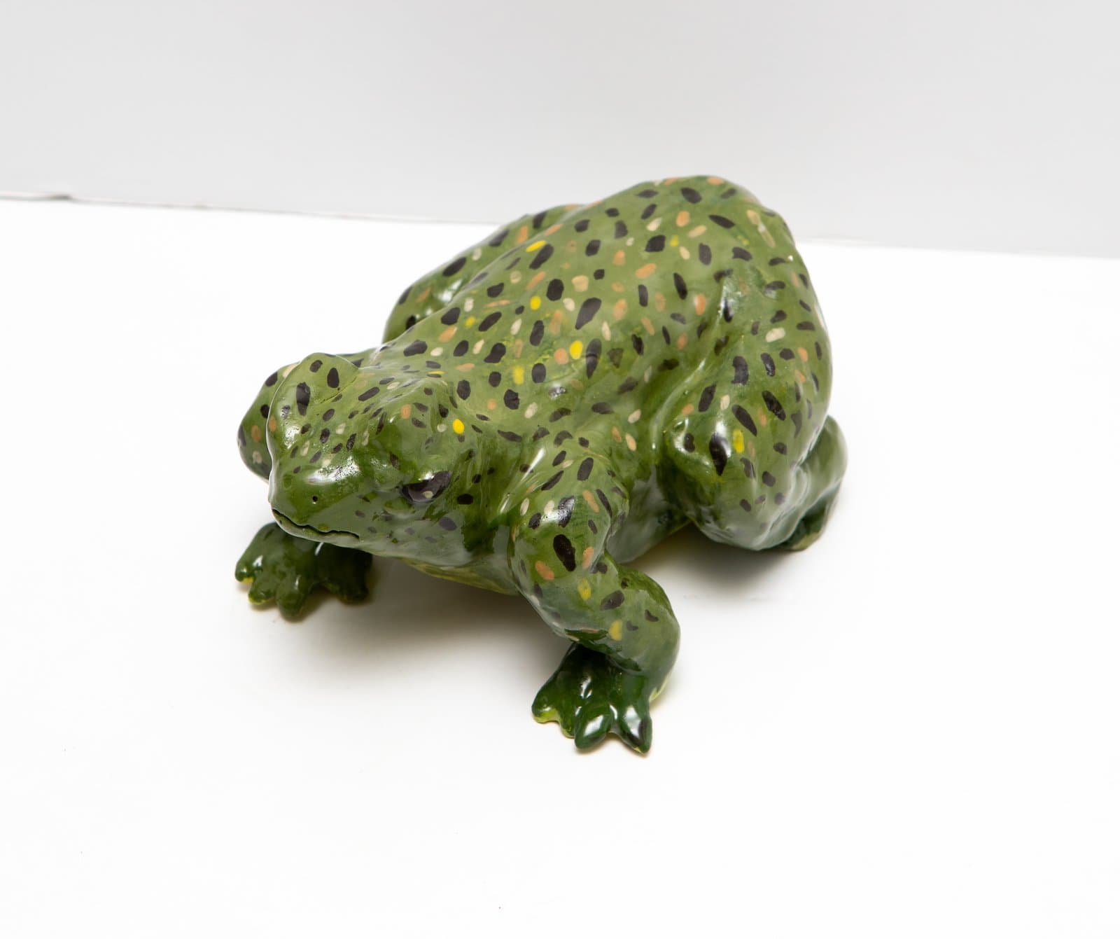 Ceramic sculpture of a squat frog in olive green glaze with multicolored spots, viewed from a three-quarter angle.