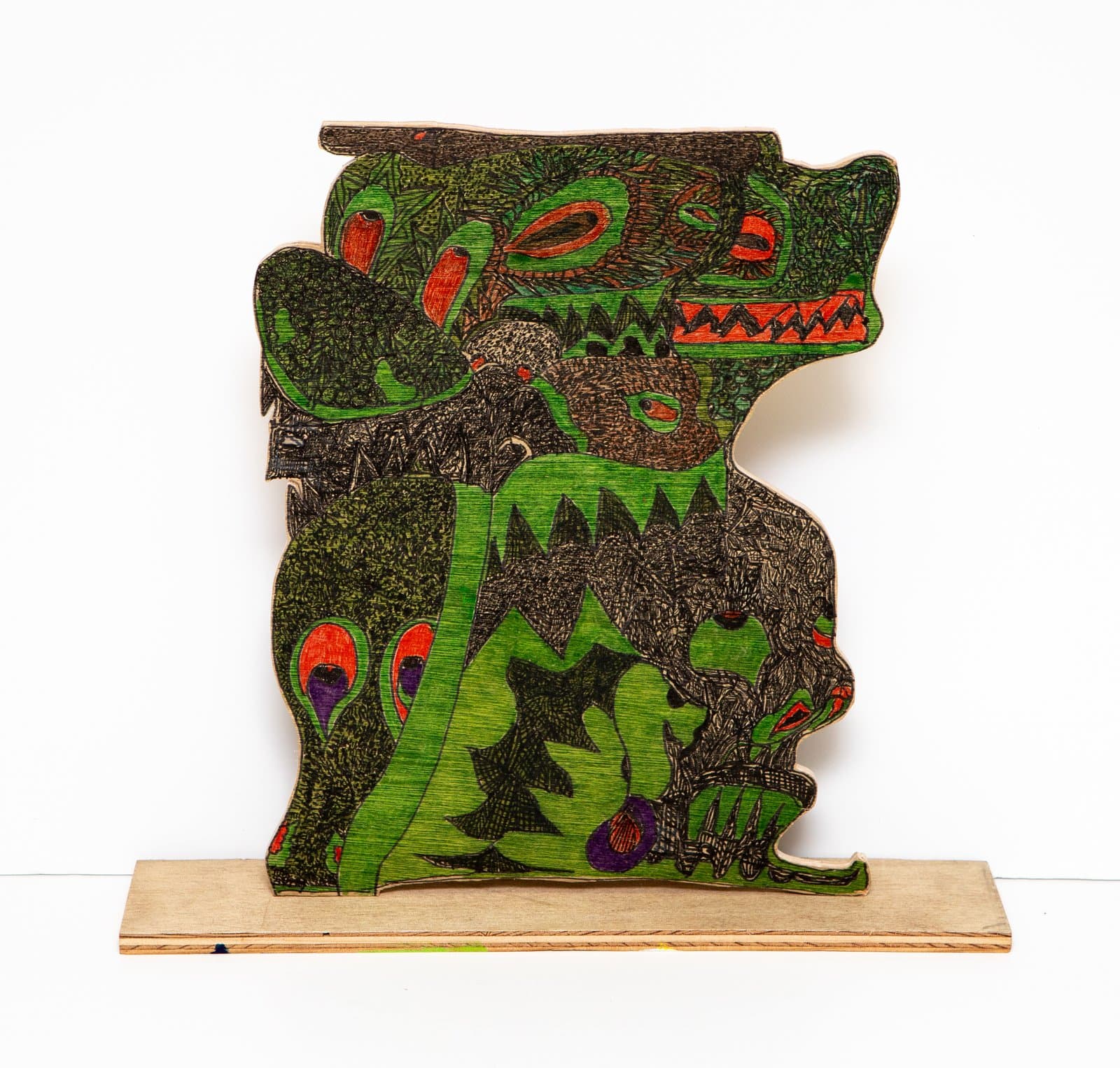 Freestanding cut wood sculpture drawn in pen, depicting a dense creature form in green, black, and red on a wood base.