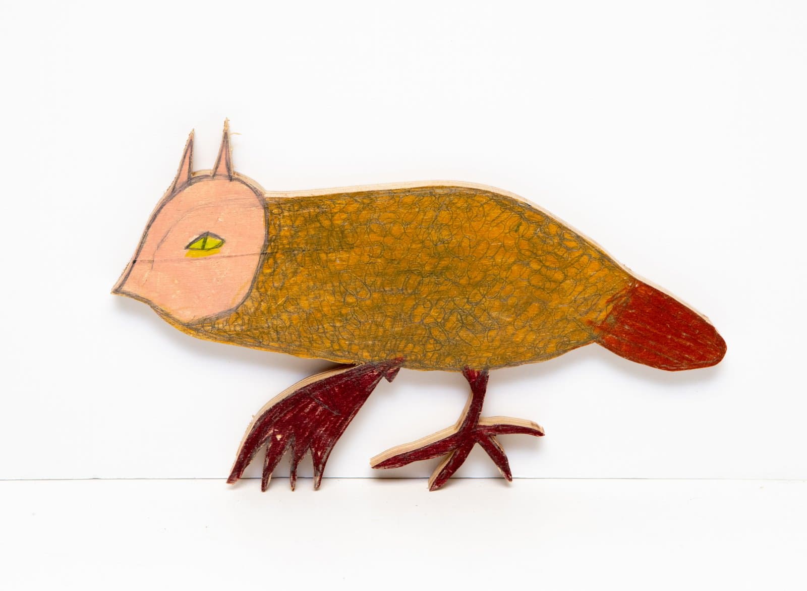 Cut wooden sculpture of a bird-like creature drawn with colored marker, featuring golden-brown body, red tail and claws, pink face.
