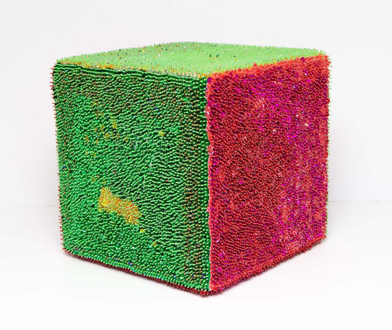 Cube sculpture densely covered in sequins, with bright green on two sides and vivid magenta-red on one visible face.