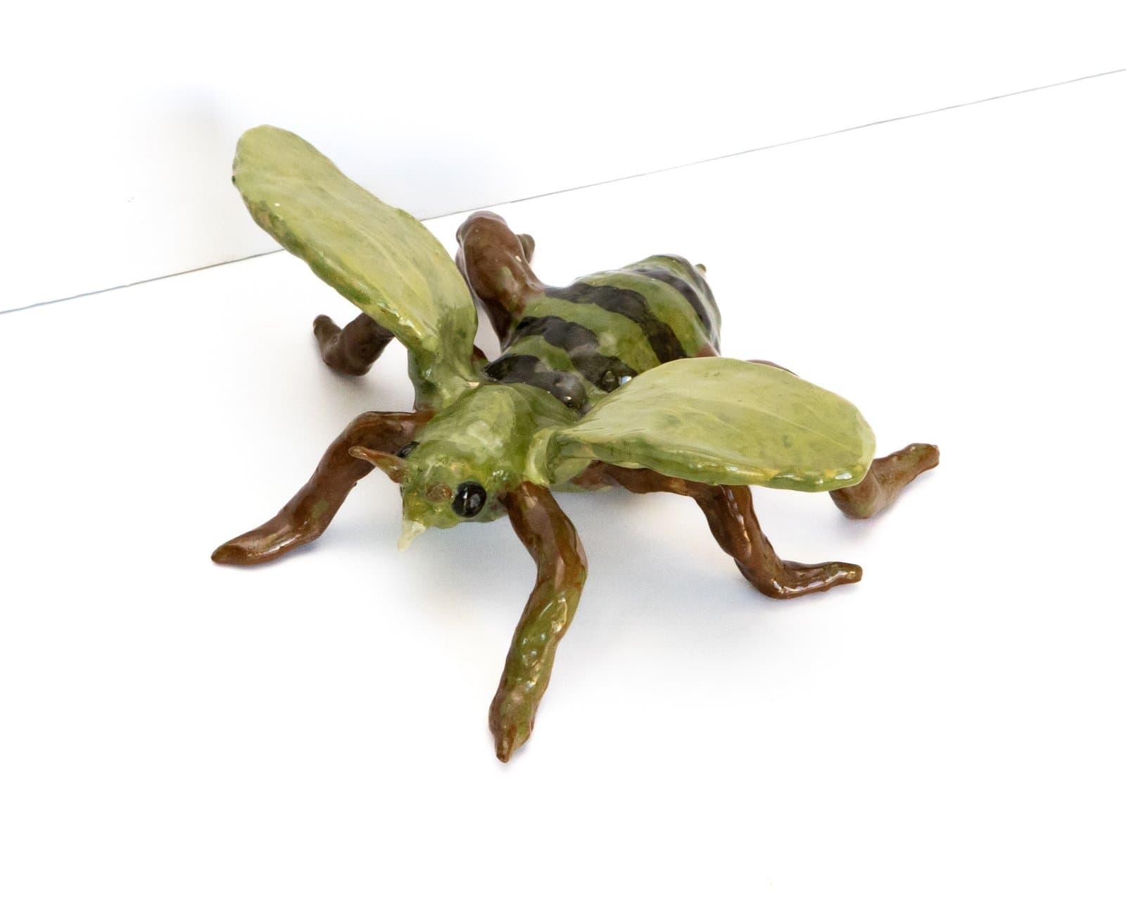 Ceramic sculpture of a bee with yellow-green wings and body, dark striped abdomen, and brown legs on white surface.
