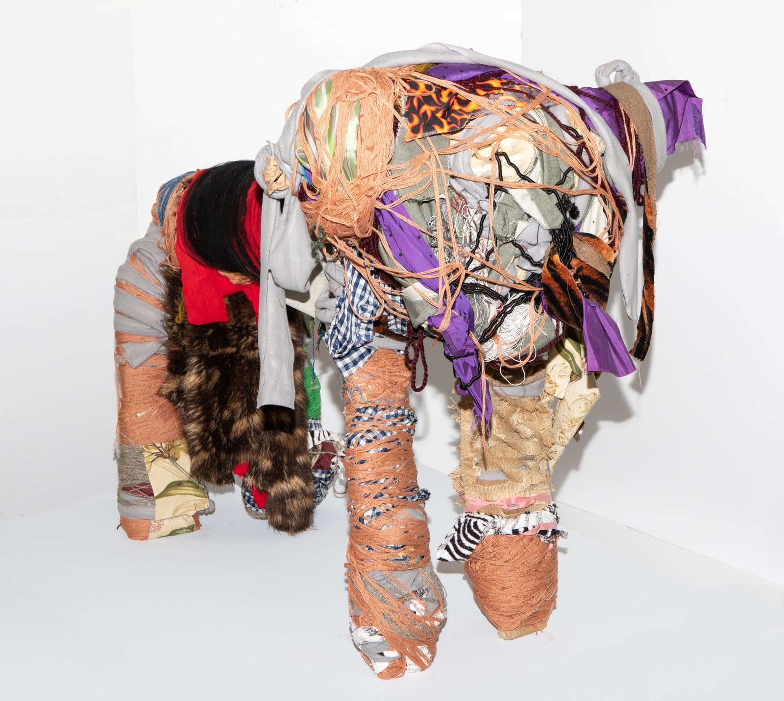 Fabric sculpture of a four-legged animal form densely wrapped in multicolored textiles, yarn, fur, and mixed materials.