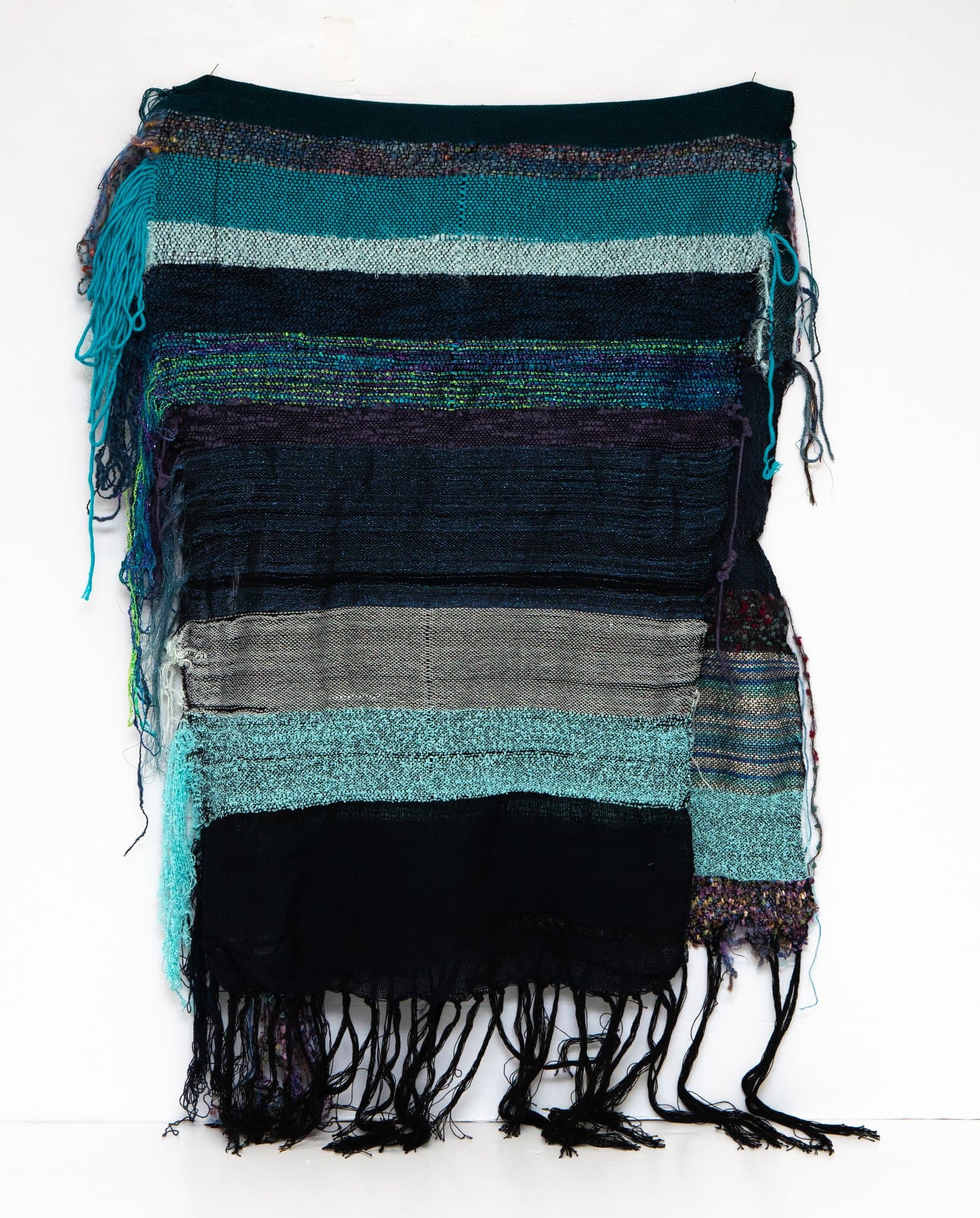 Woven textile wall hanging with horizontal stripes in teal, black, gray, and purple, with fringe along the sides and bottom.