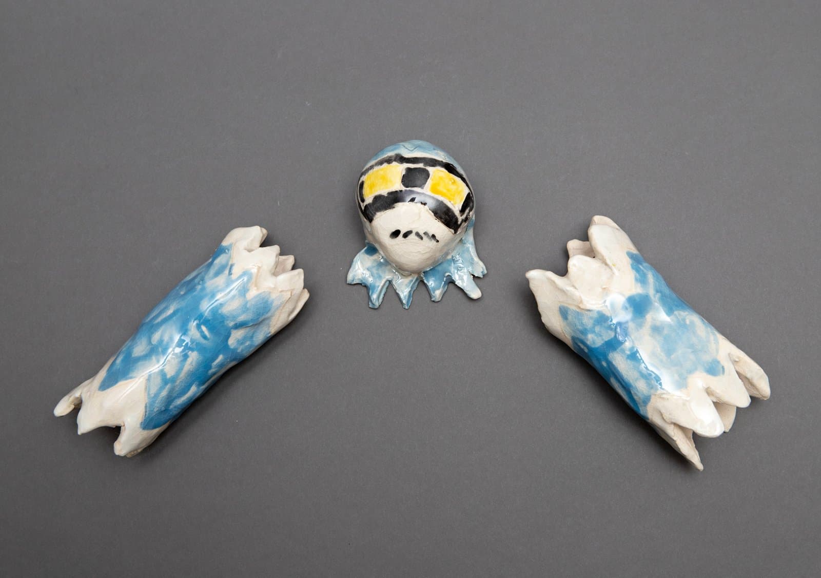 Three-piece glazed ceramic sculpture: a round creature head flanked by two wing-like forms in blue, white, yellow, and black.