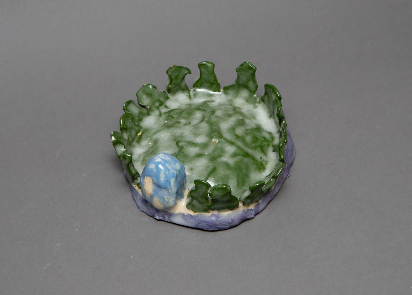 Small round ceramic dish with jagged green rim, mottled green and white interior, and a blue oval form on the side.