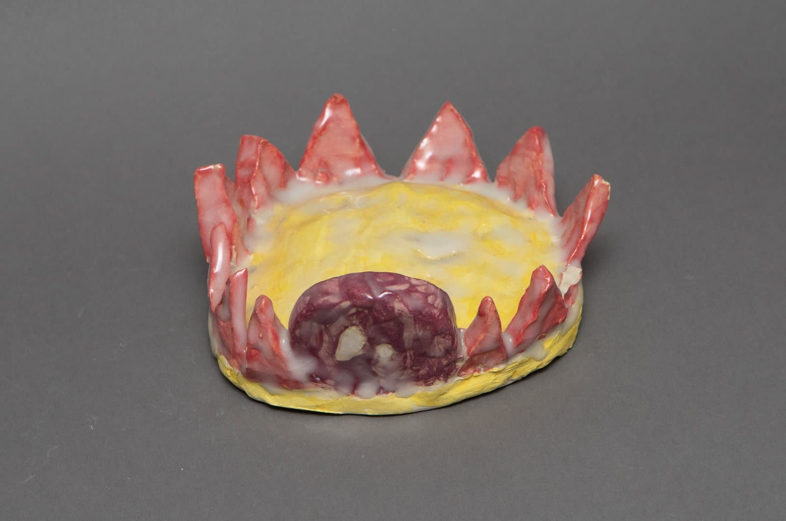 Glazed ceramic sculpture resembling a low crown or sun form with red pointed spikes, yellow body, and purple central mound.