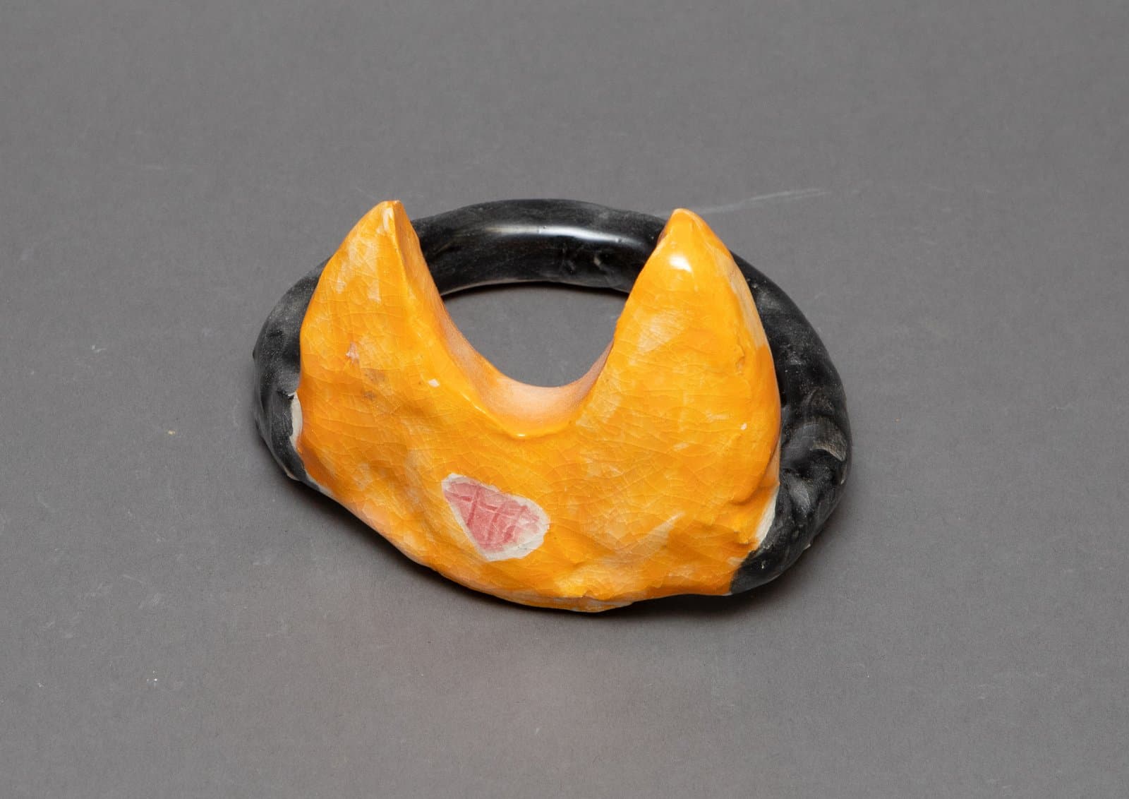 Small ceramic sculpture with a black ring base and orange double-peaked form, featuring a small pink and white accent.