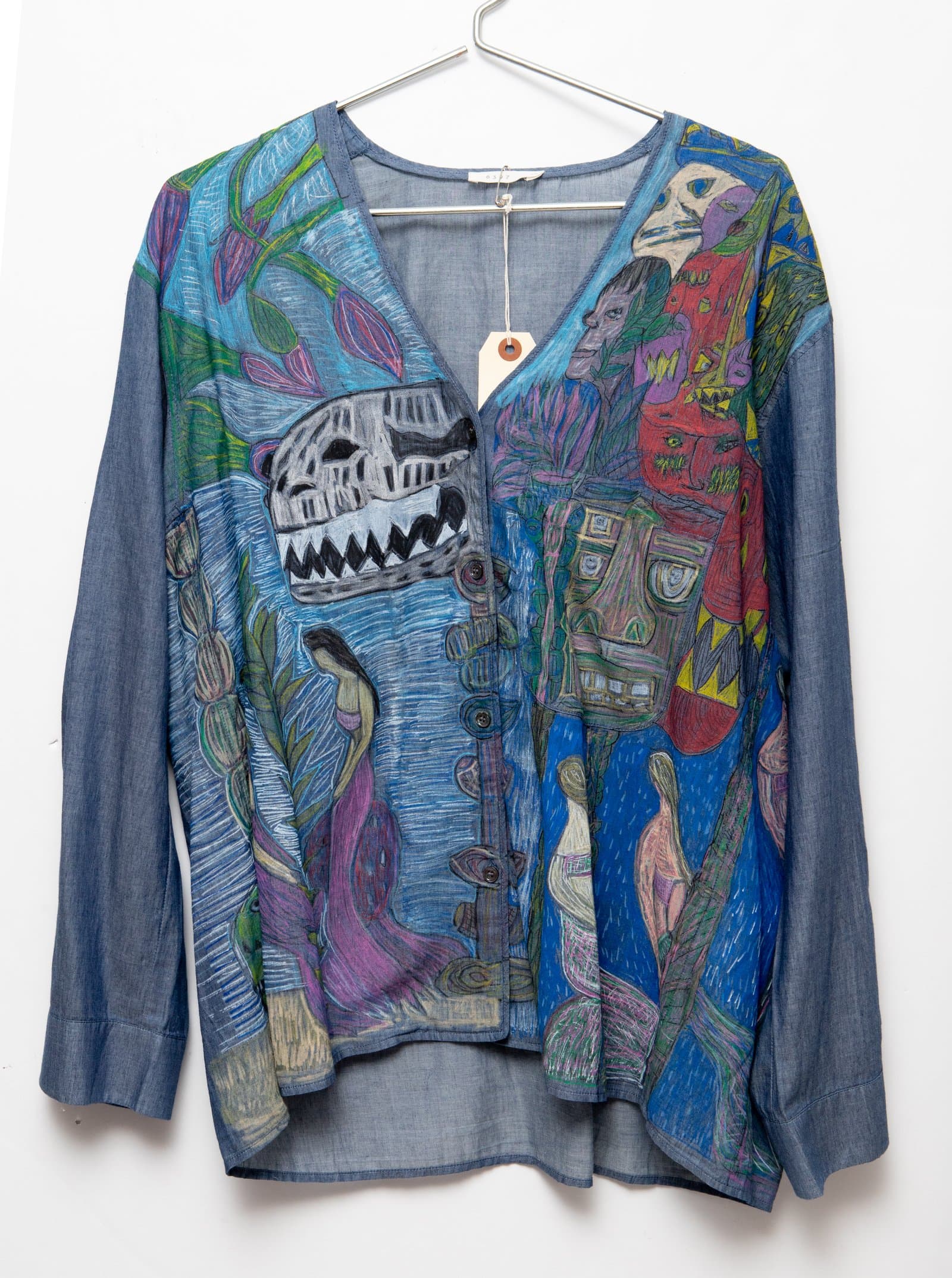 Light blue chambray button-front shirt hand-drawn with dense figurative imagery including masked faces, human figures, and botanical forms.