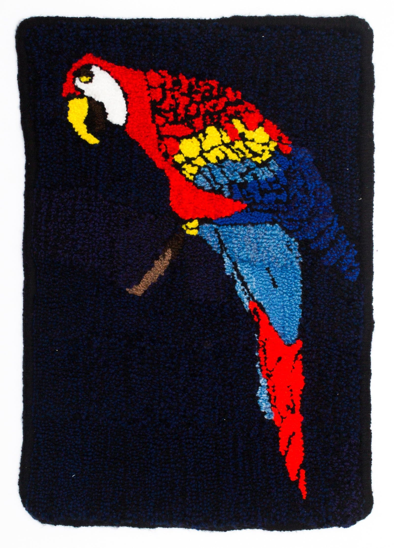 Latch hook textile depicting a scarlet macaw perched on a branch, rendered in red, blue, yellow, and white yarn on a dark navy background.