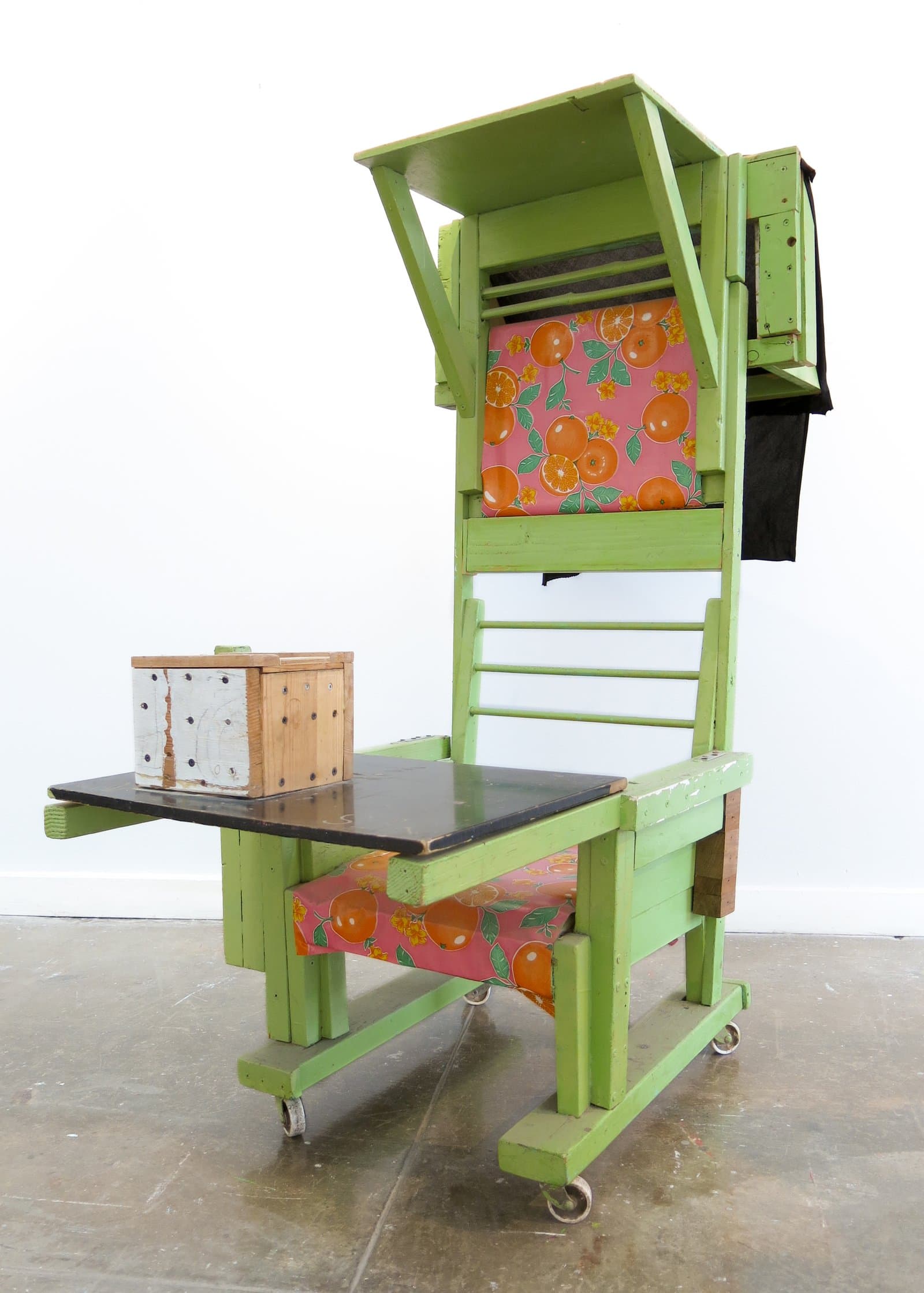Tall sculptural chair painted bright green with orange-print fabric cushions, a side tray, and caster wheels.