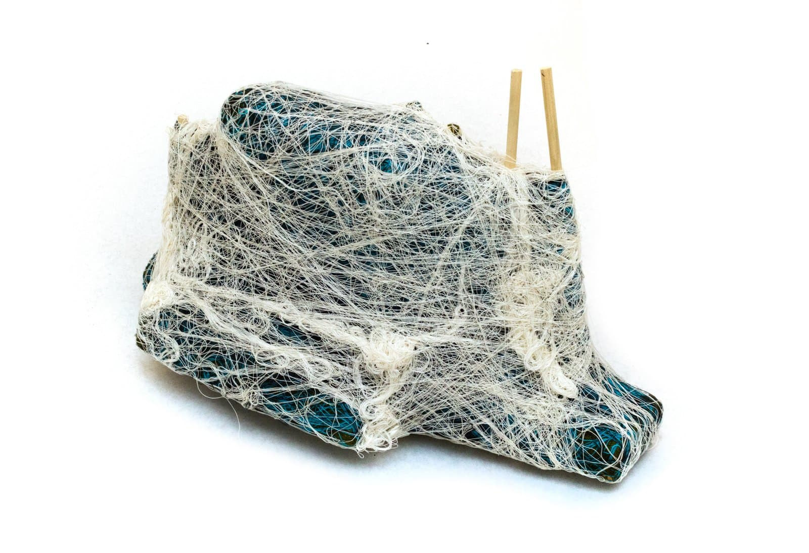 Mixed-media sculpture of a wrapped object densely covered in white fiber strands with teal and black fabric visible beneath, two wooden sticks protruding from top.