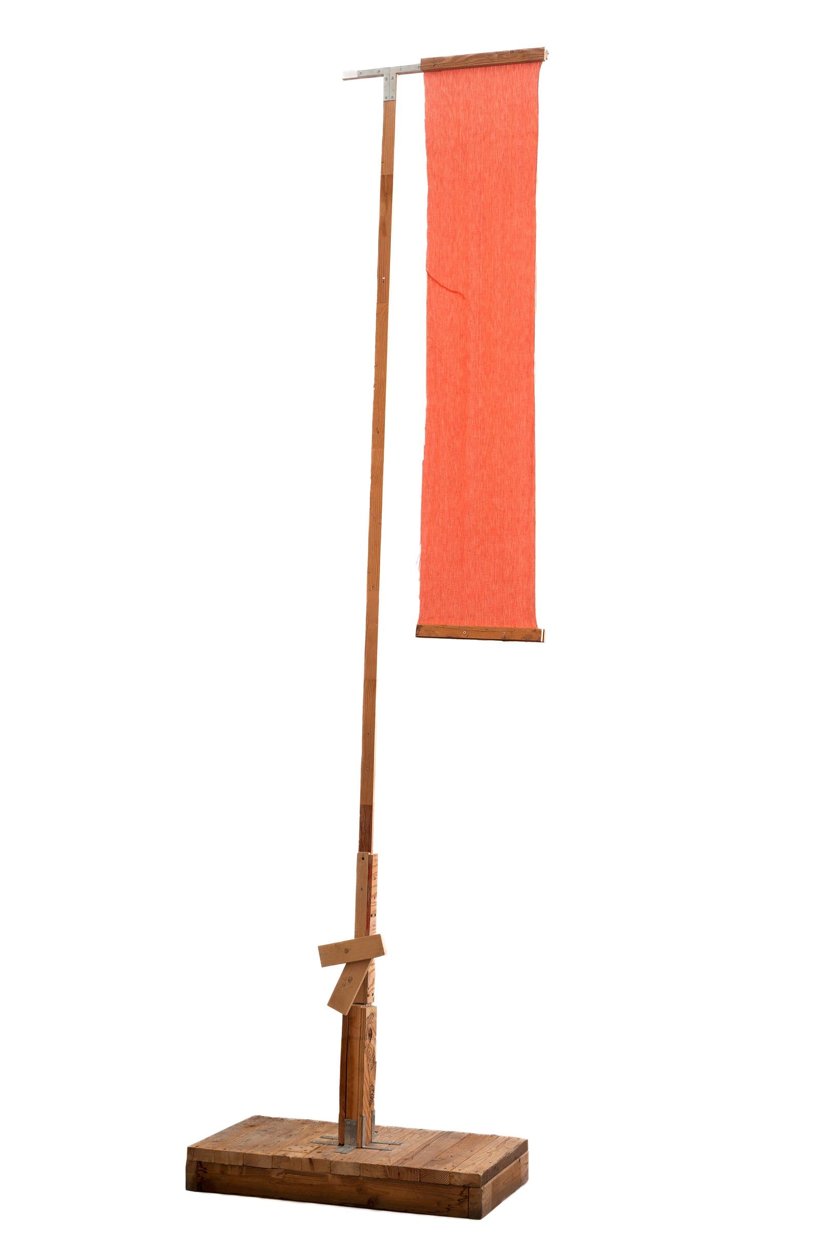 Tall freestanding sculpture with a wooden pole and base suspending a vertical coral-orange textile banner via a metal crossbar.