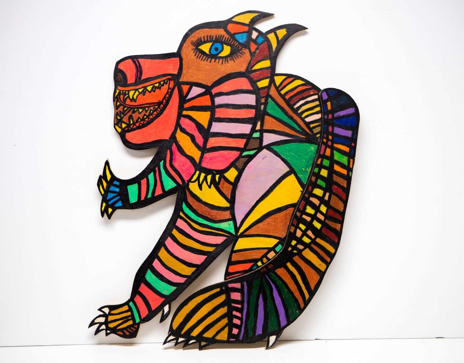 Cutout painted wood silhouette of a fanged, upright creature covered in bold multicolored stripes and geometric sections.