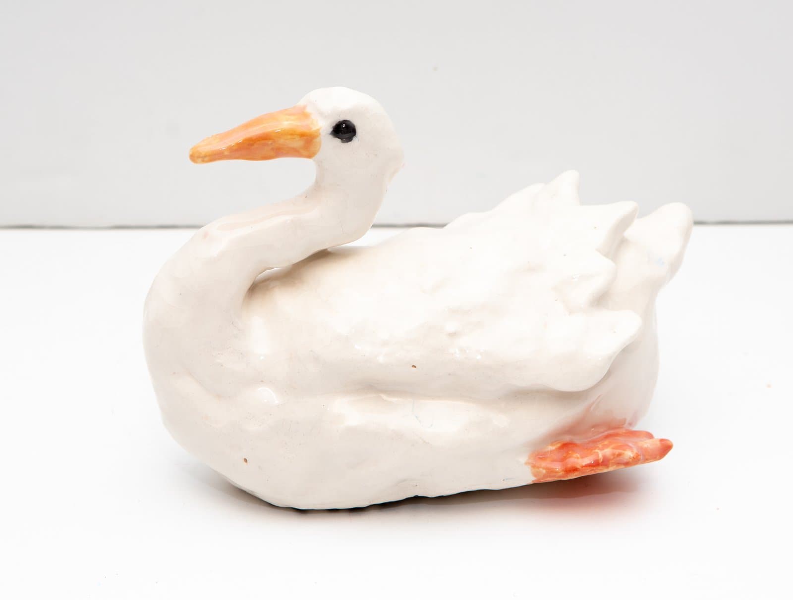 Ceramic sculpture of a seated white goose with an orange beak, black eye, and orange-red webbed feet.