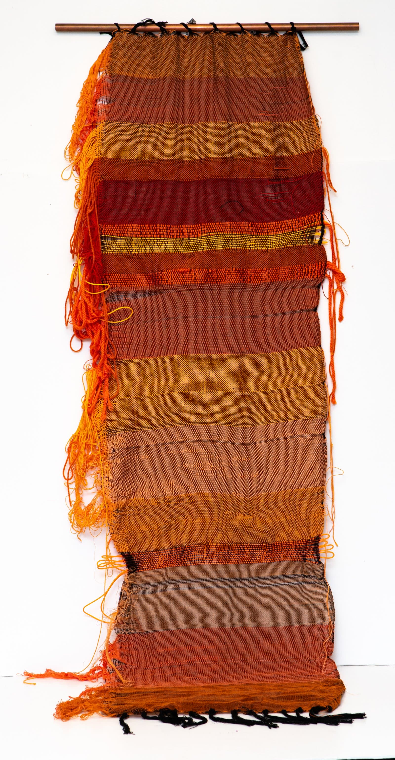 Woven textile wall hanging in horizontal bands of orange, rust, brown, and gold, suspended from a copper rod.