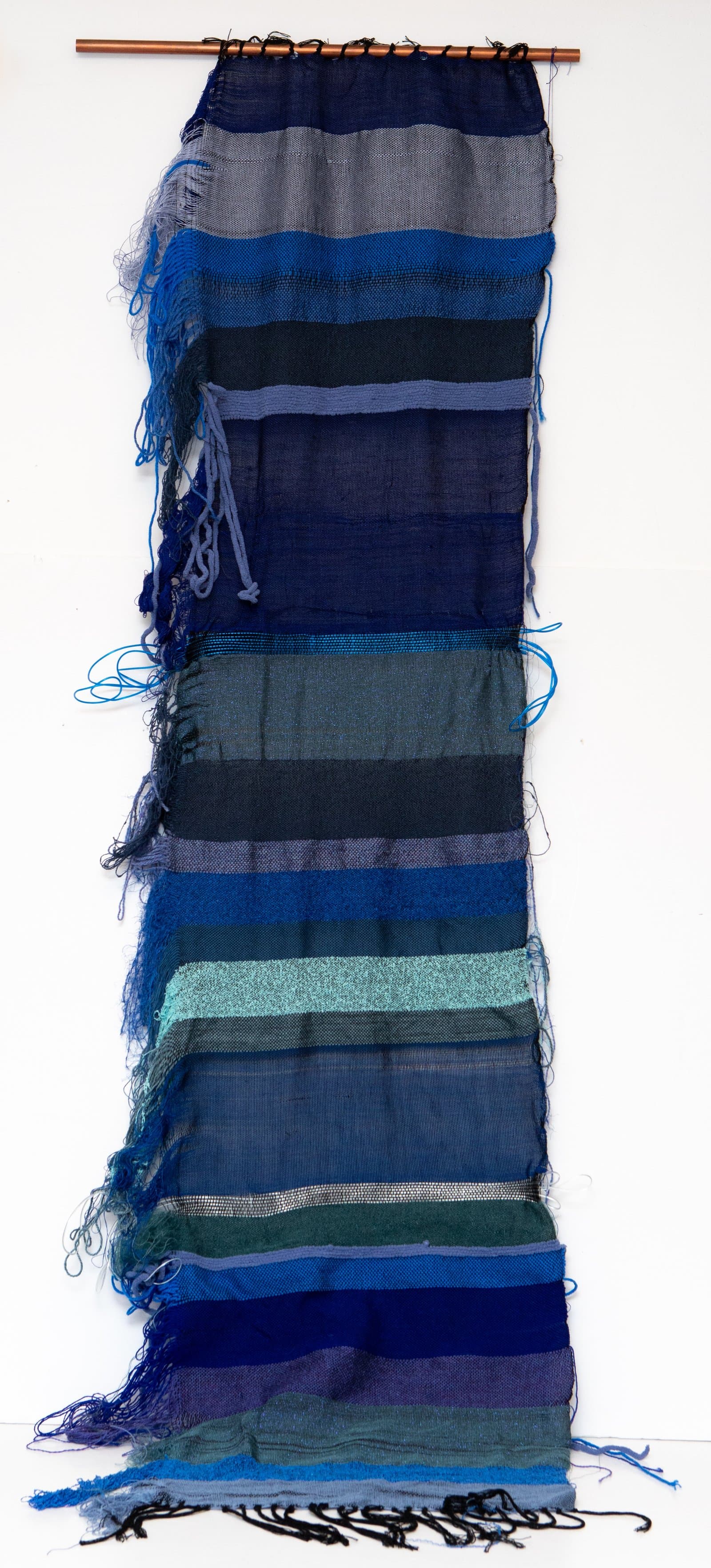 Tall woven textile wall hanging in horizontal stripes of blue, teal, navy, and gray, suspended from a copper rod.