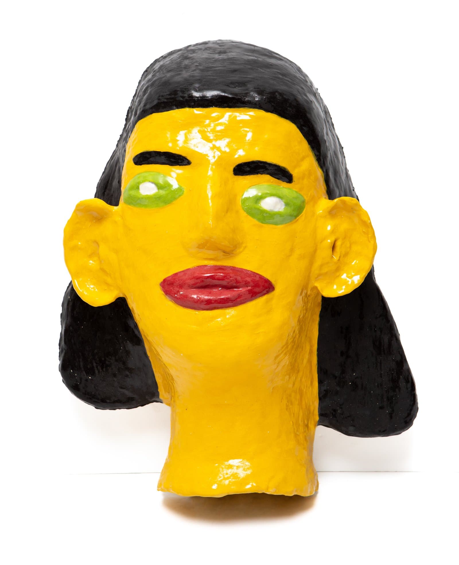 Ceramic sculpture of a human head with bright yellow glaze, black hair, green eyes, and red lips on white background.