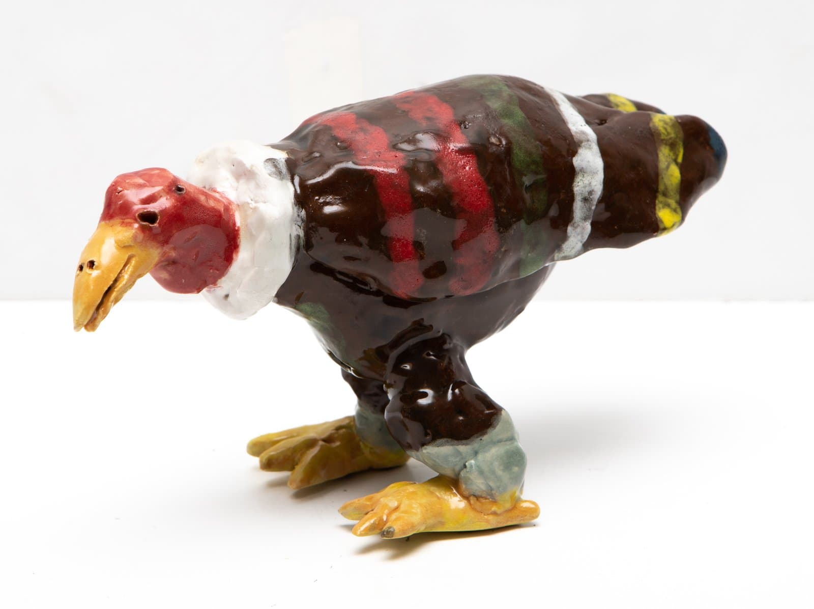 Glazed ceramic sculpture of a bird with dark brown body, red head, yellow beak, and multicolored accents on white surface.
