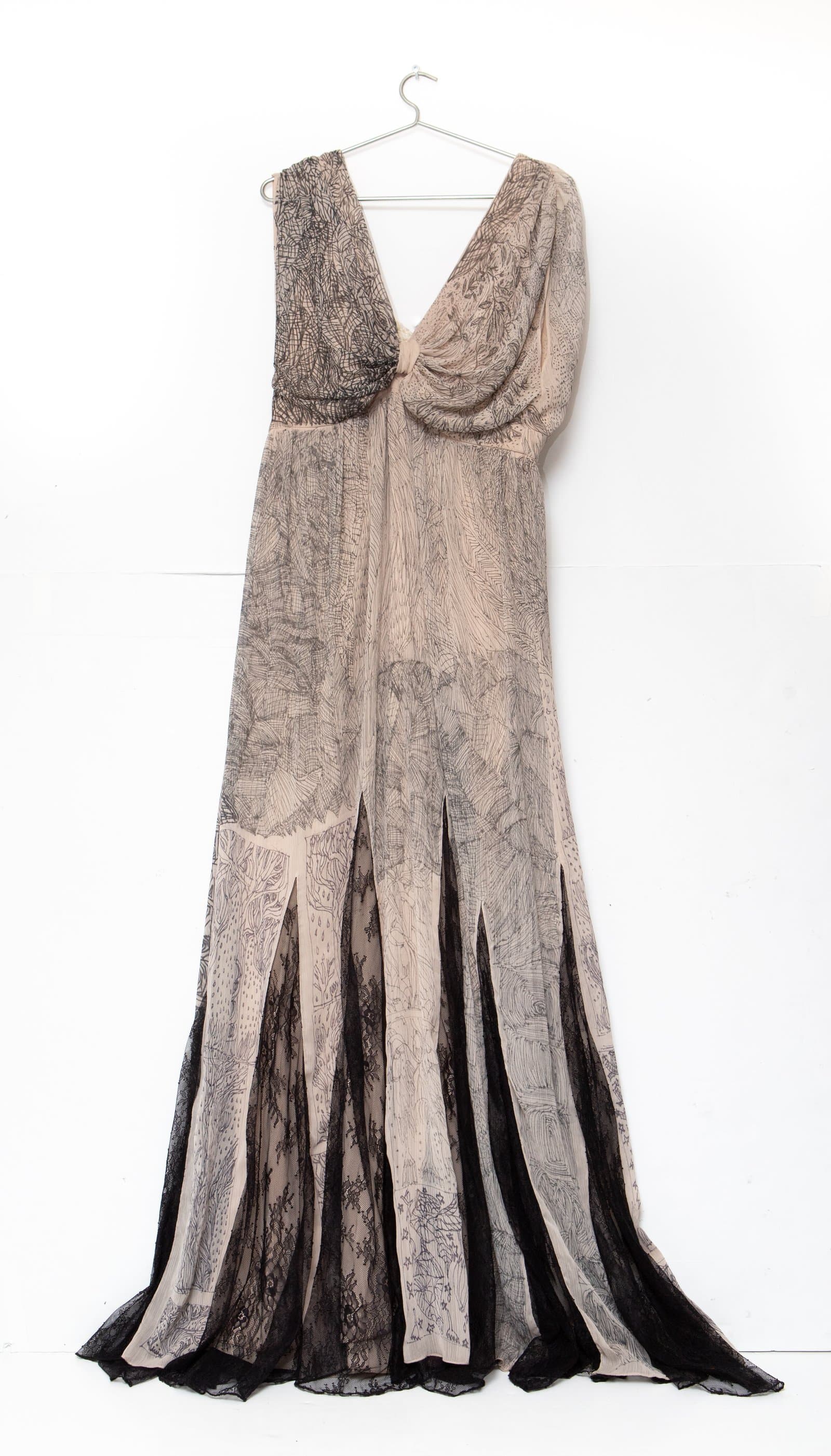 Floor-length dress on a wire hanger, covered in dense black ink drawings on cream fabric with black lace trim at hem.