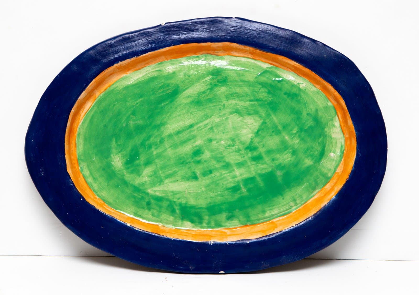 Oval ceramic plate with concentric bands of navy blue, orange, and green; brush marks visible on green center.