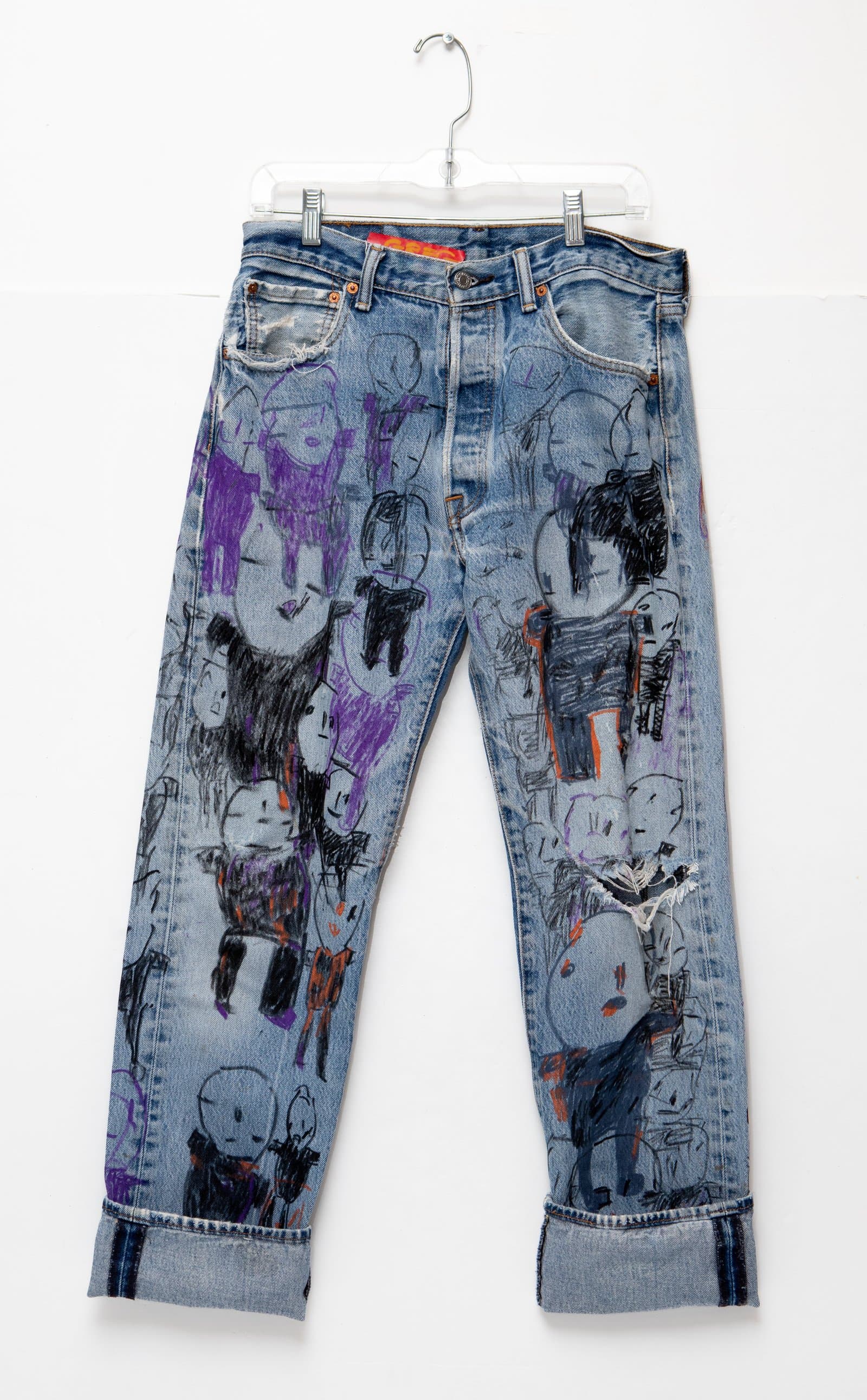 Light-wash denim jeans covered with hand-drawn figures in black, purple, and orange marker, displayed on a hanger.