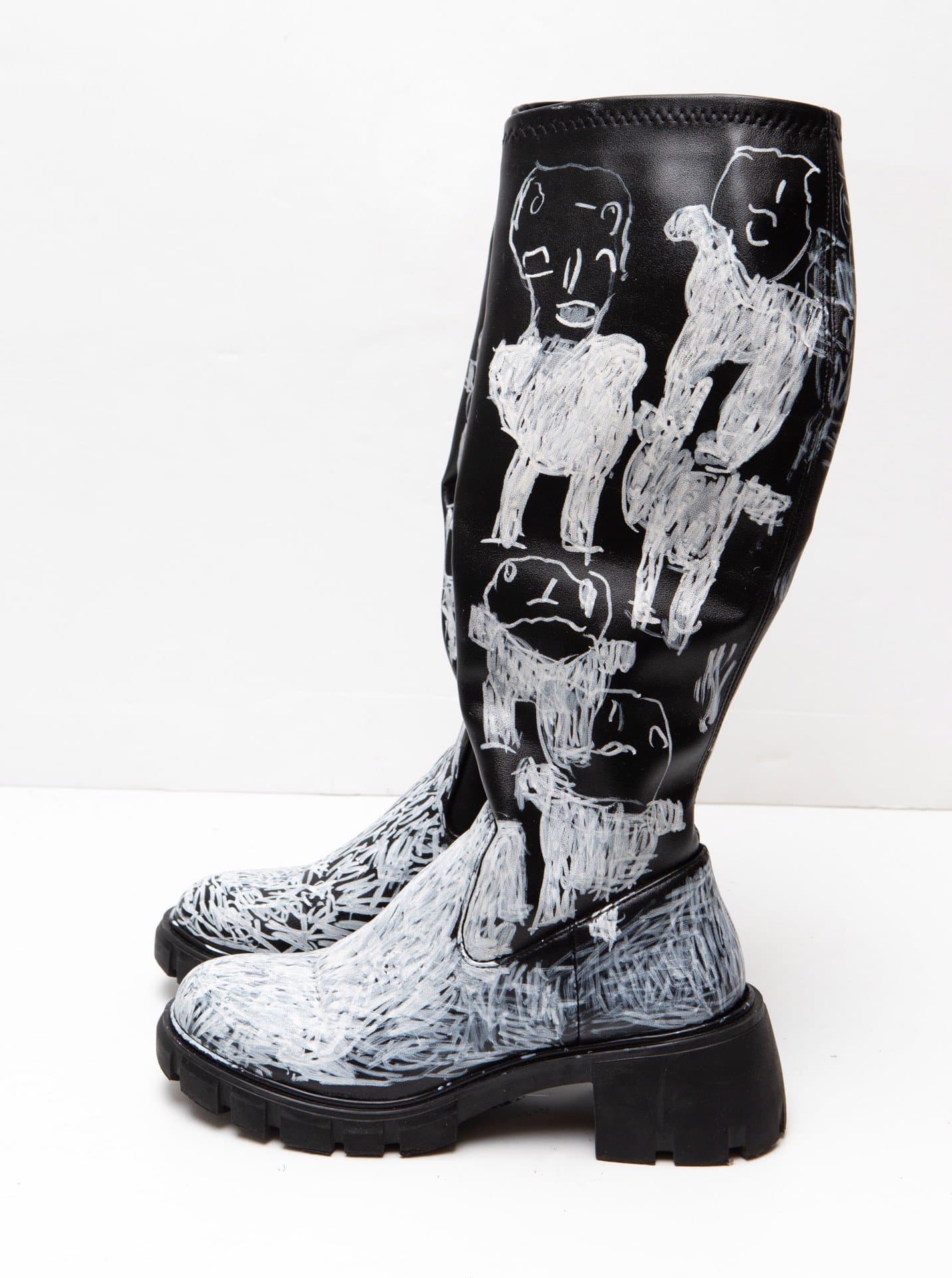 Decorated black leather boot with white chalk or paint drawings of multiple human figures on dark surface.