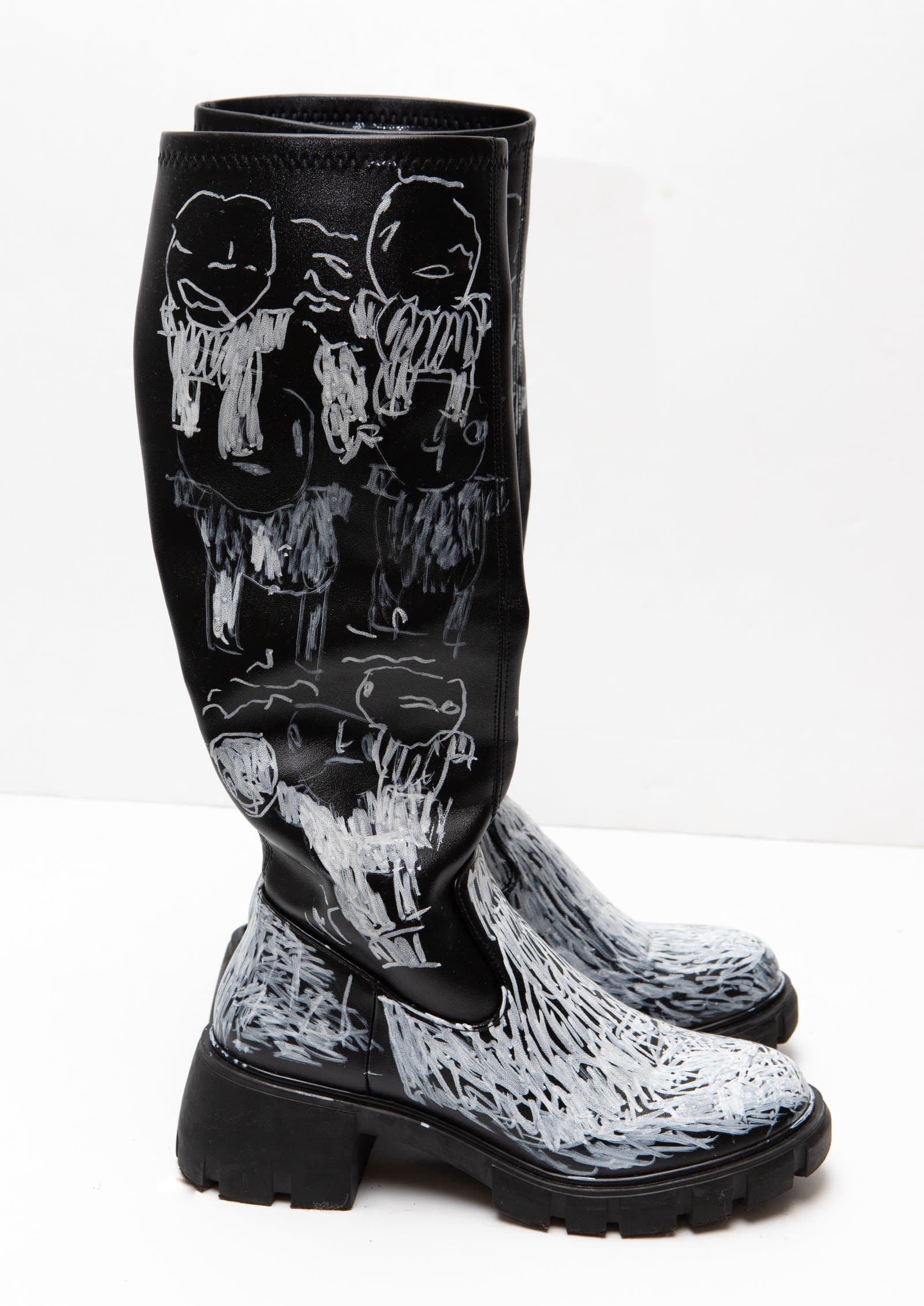 Pair of black knee-high boots hand-drawn with white marker depicting human figures on the shaft and dense hatching on the toe.