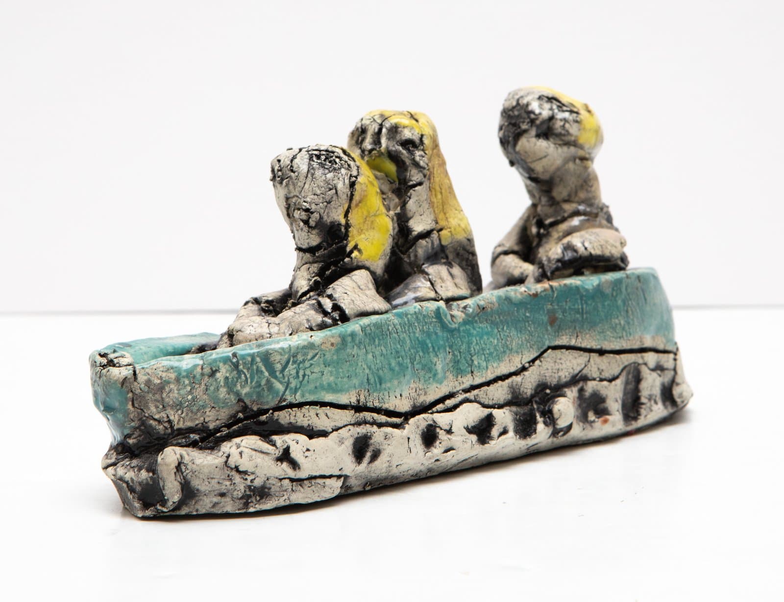 Ceramic sculpture of three figures seated in a turquoise boat, with yellow hair accents and a heavily textured gray-black surface.