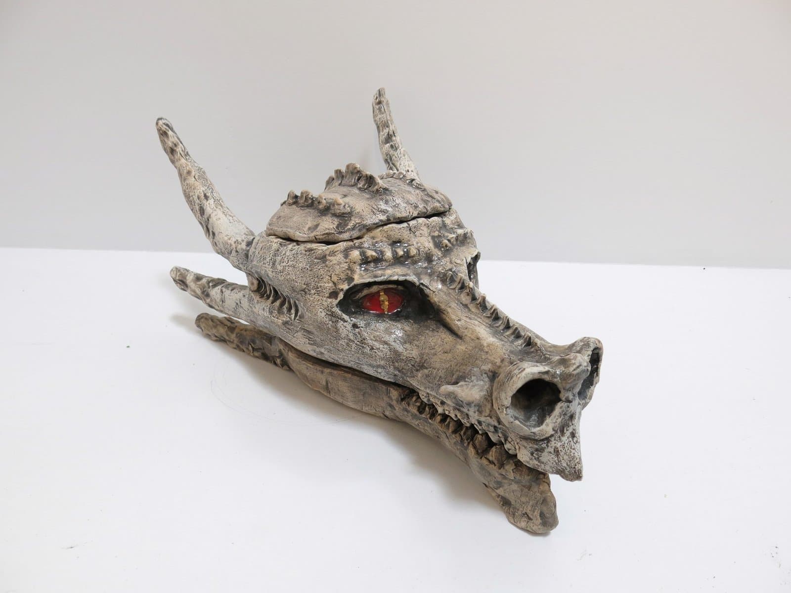 Sculptural dragon head mask in weathered gray ceramic with horns, teeth, and red glass eye detail