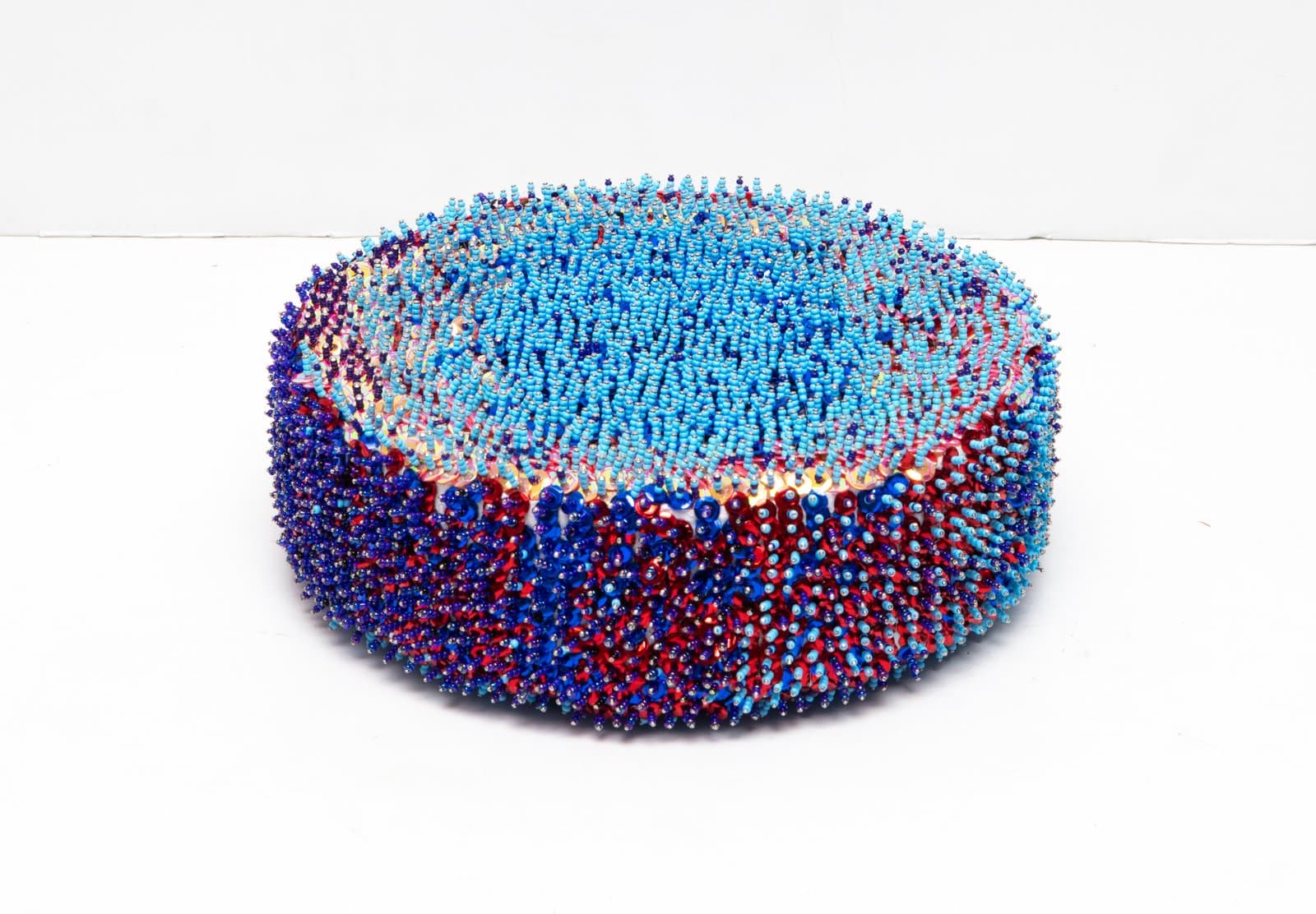 Circular drum-shaped sculpture densely covered in blue, purple, and red sequins or beads against a white background.