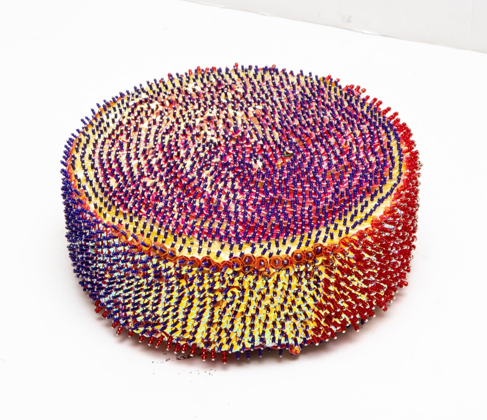 Cylindrical sequin sculpture densely covered in blue, red, and yellow sequins arranged in radiating and swirling patterns.