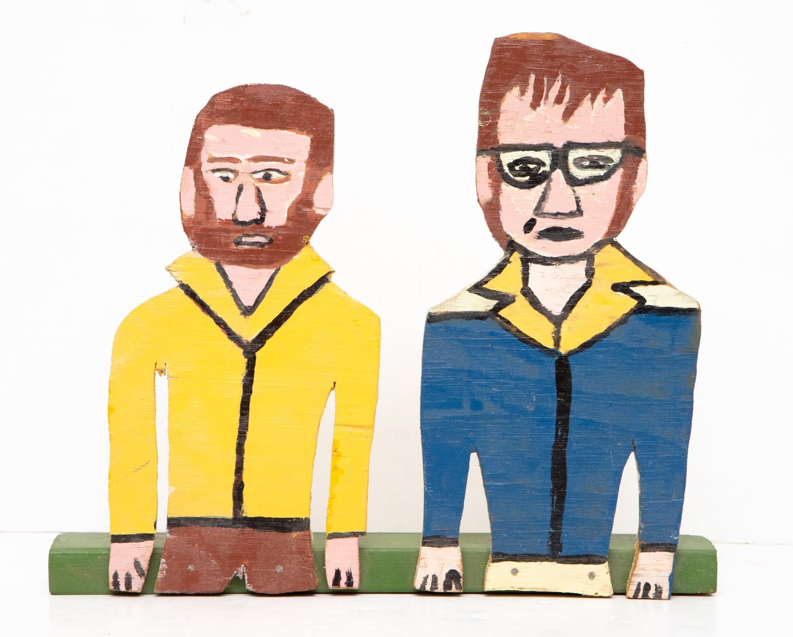 Painted wood sculpture of two standing figures in yellow and blue jackets mounted on a green base.