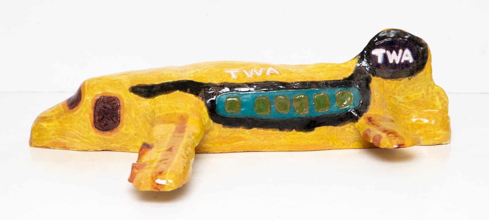 Glazed ceramic sculpture of a reclining yellow dog with "TWA" lettering, teal windows, and a black stripe along its body.