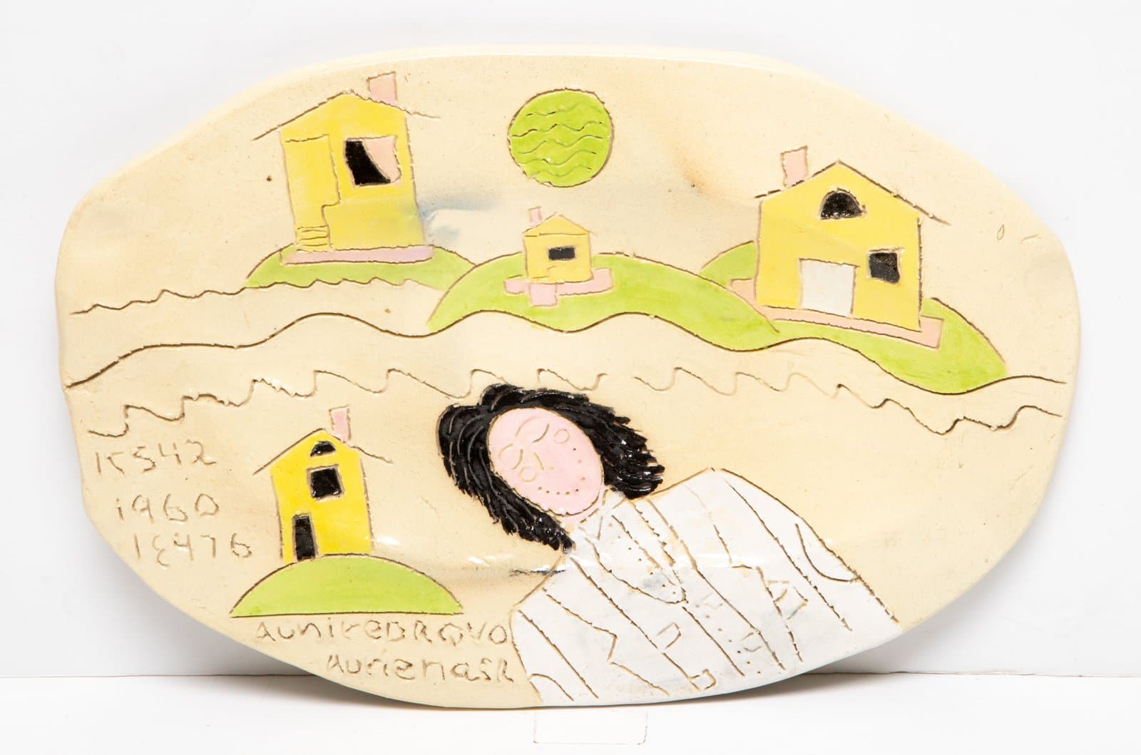 Oval ceramic relief plaque with yellow houses on green hills, a reclining figure with black hair, and inscribed text.