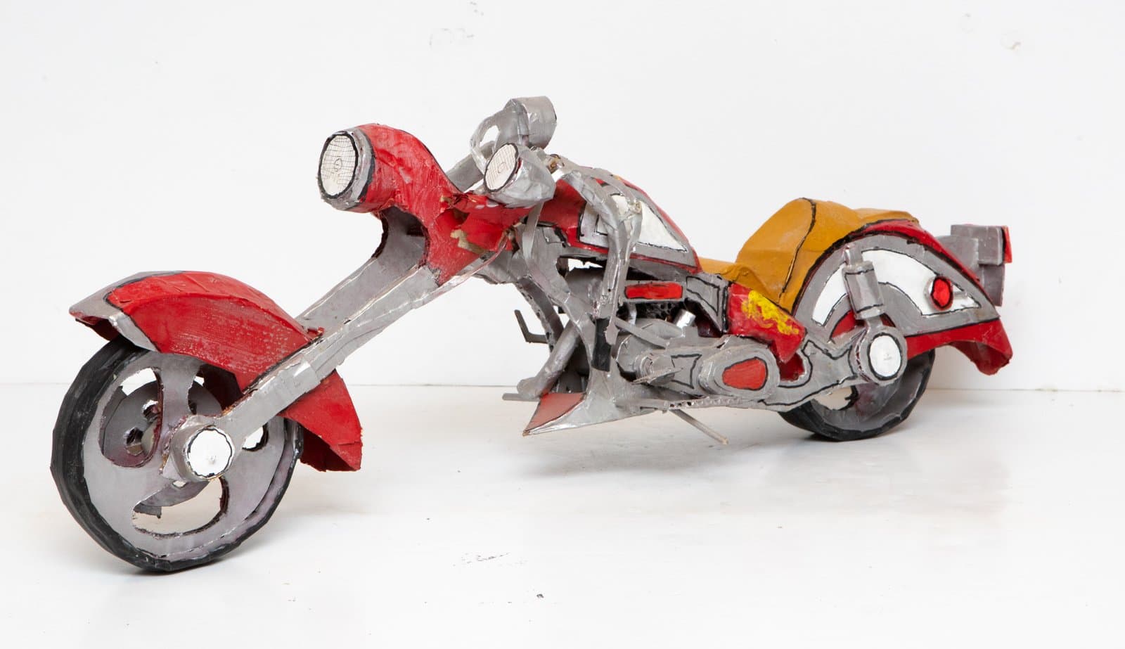 Handmade cardboard sculpture of a chopper-style motorcycle painted in red, silver-gray, and mustard yellow on white background.